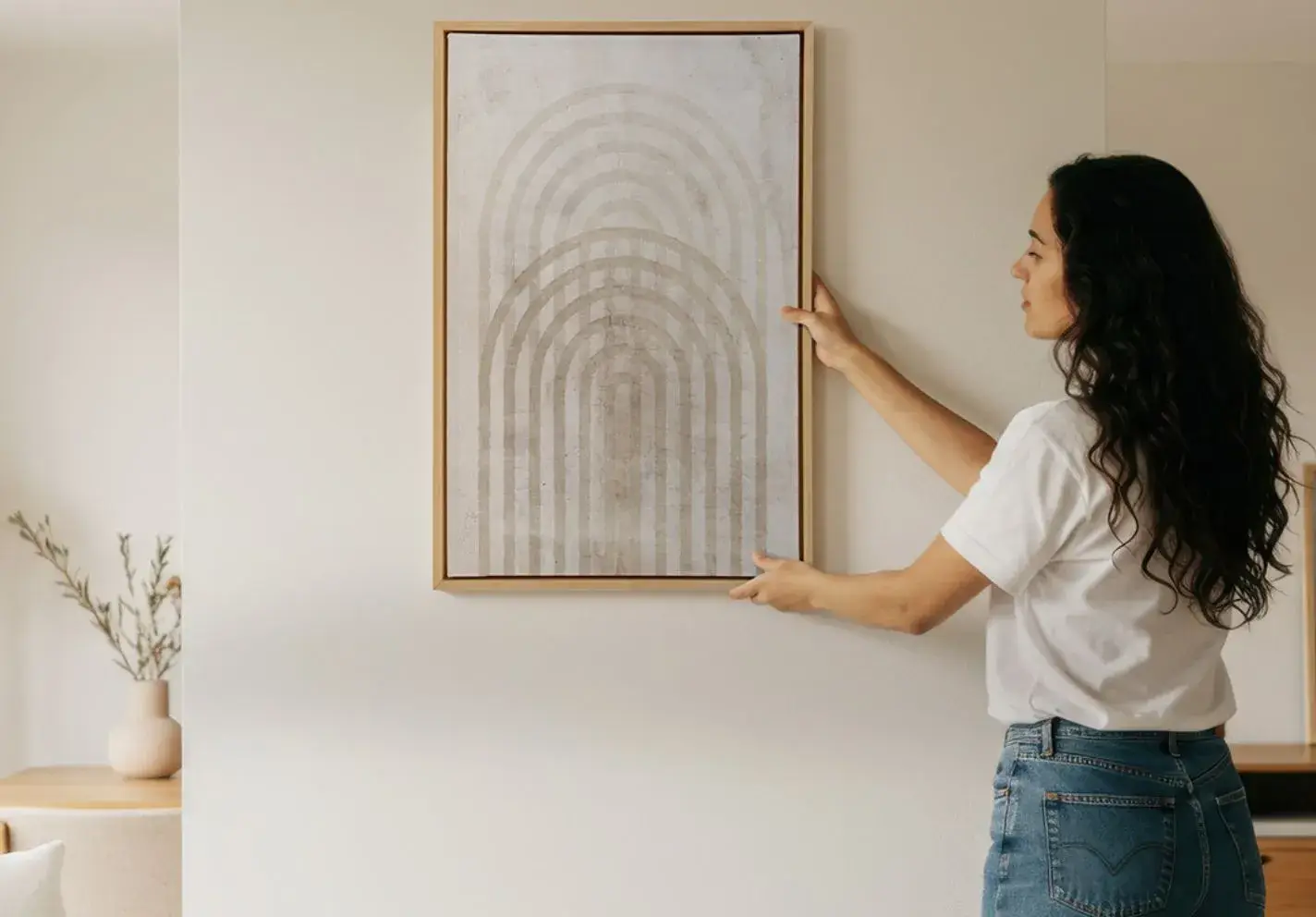 The woman hangs the painting on a light-coloured wall. The painting, in a wooden frame, features a minimalist design with a motif of vertical, overlapping arches in muted shades of beige and grey.